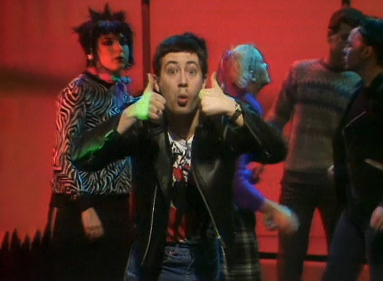host Baz giving a thumbs up, surrounded by young adults dancing
