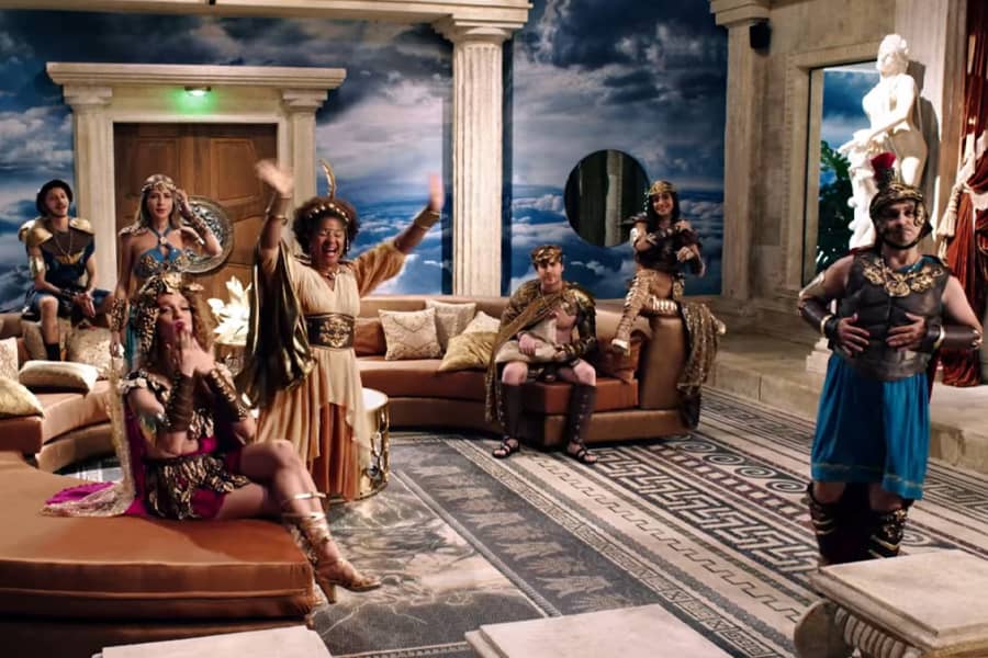 the Greek gods in a fancy living room waving at the audience