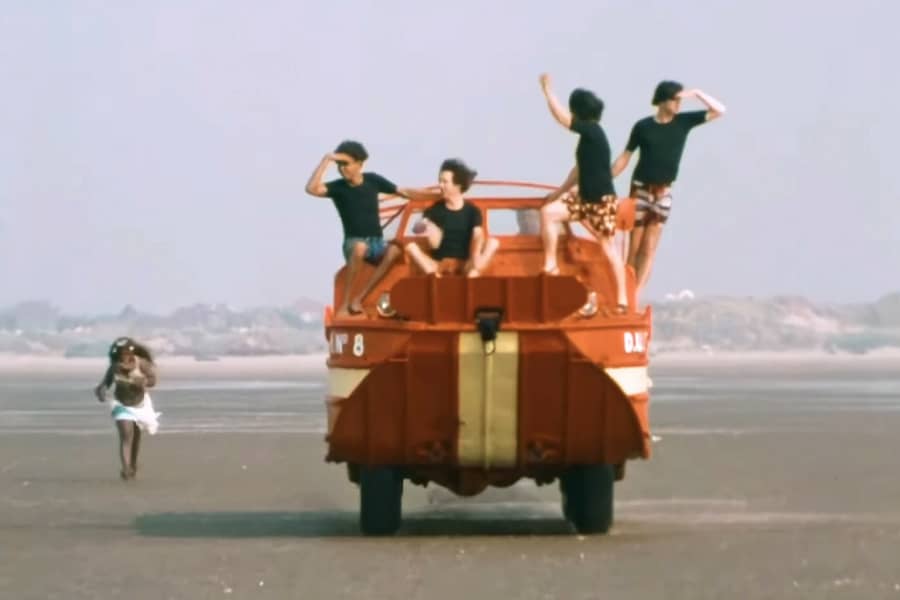 the Rutles riding a beach boat car while a native woman runs after them