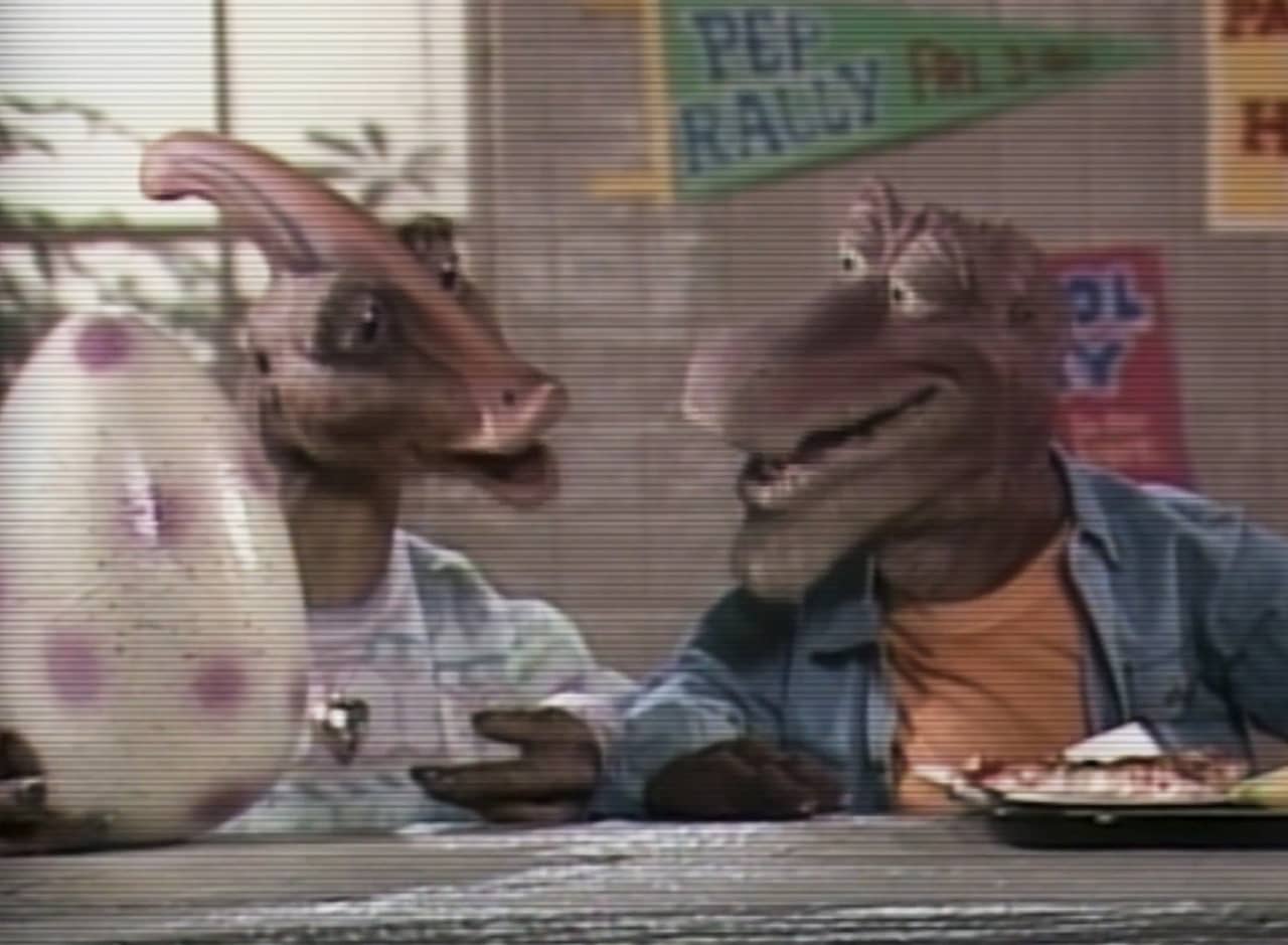 a teen boy and girl talking in the cafeteria, the girl has a dinosaur egg