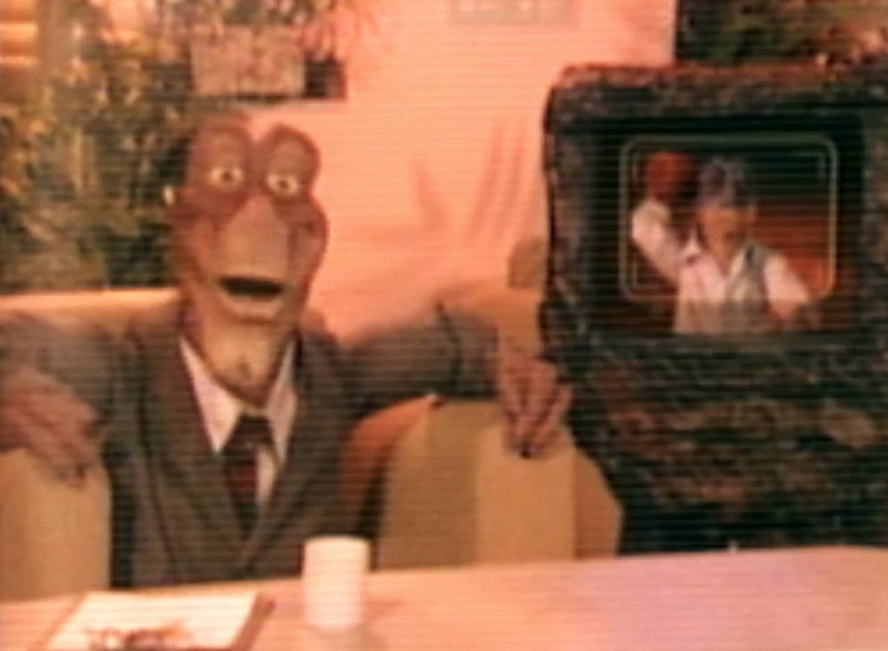 host of the show next to a TV monitor showing a guy by a gurgling volcano