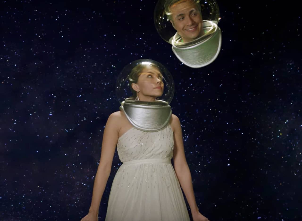 a field of stars with a woman in a fancy dress and space helmet looking up at Rogelio De La Vega’s floating head (also in a space helmet)