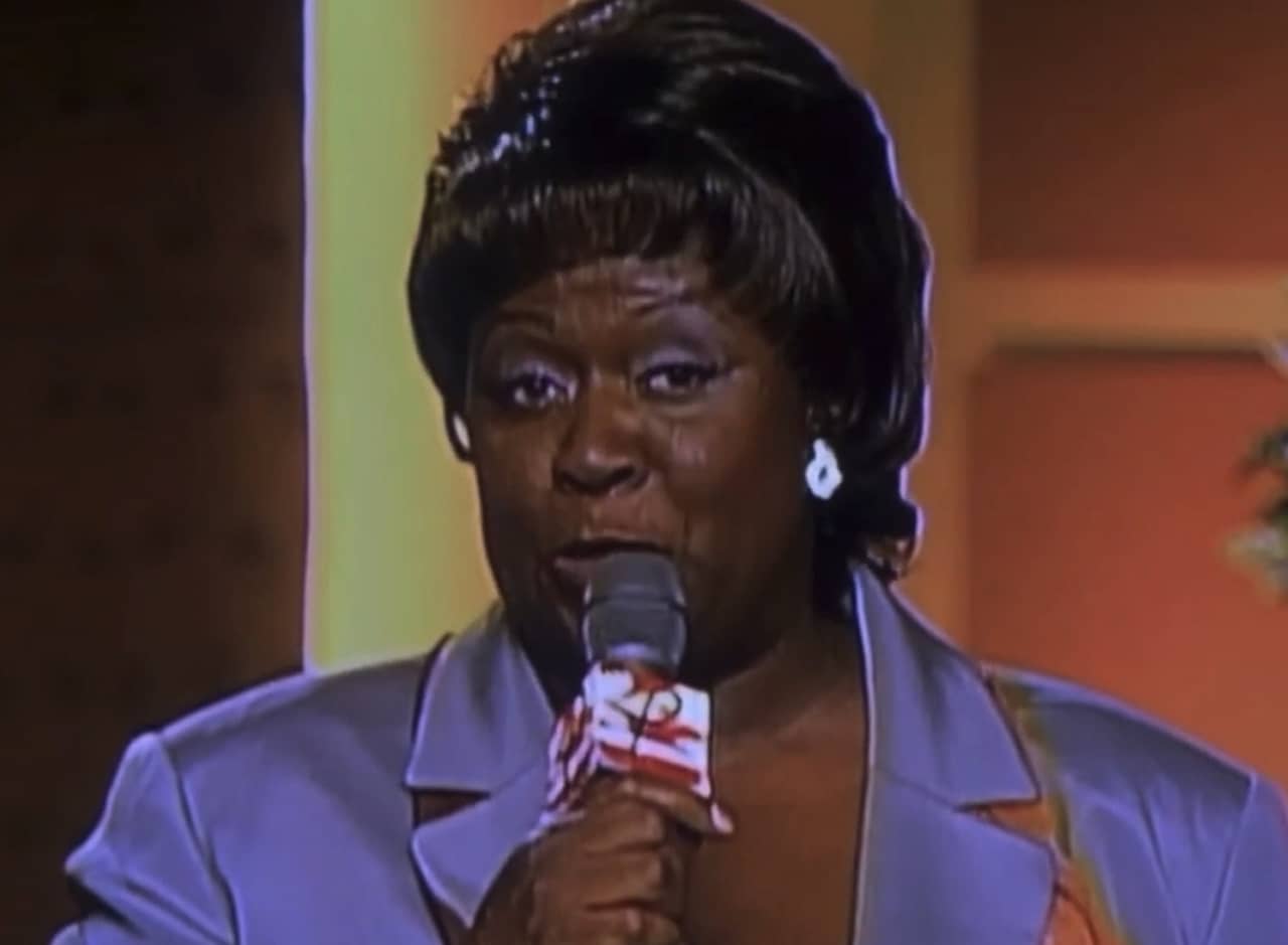 Pauletta, a black woman with short hair wearing pastel purple, speaks into a microphone on set