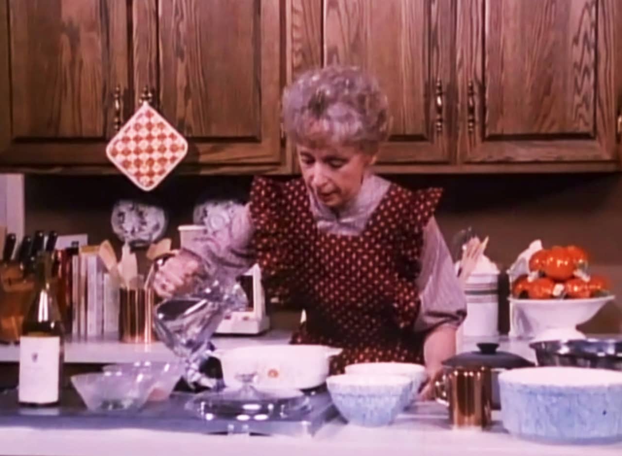 Pearl, an older woman wearing a ruffly apron, cooking in a cozy kitchen