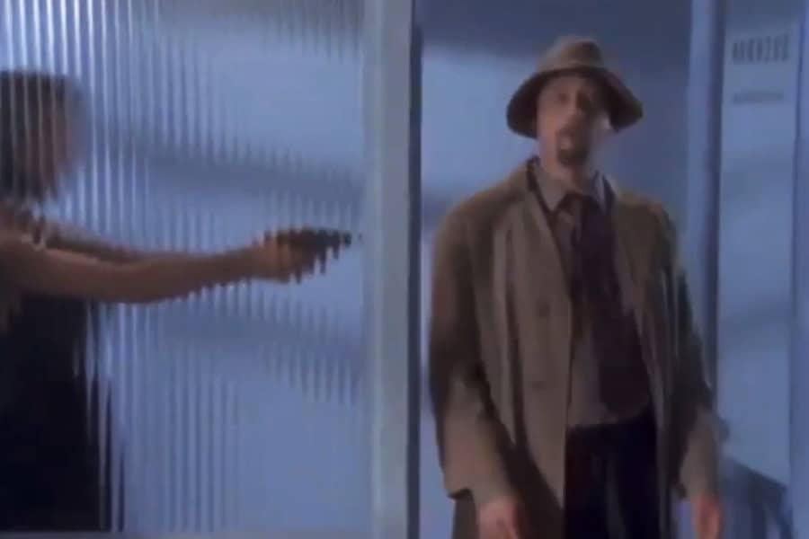 man in a trench entering the office, behind reeded glass window is a woman pointing a gun at him