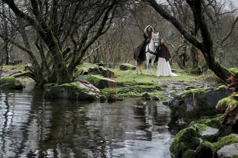 two women wait on horseback in the woods near a stream