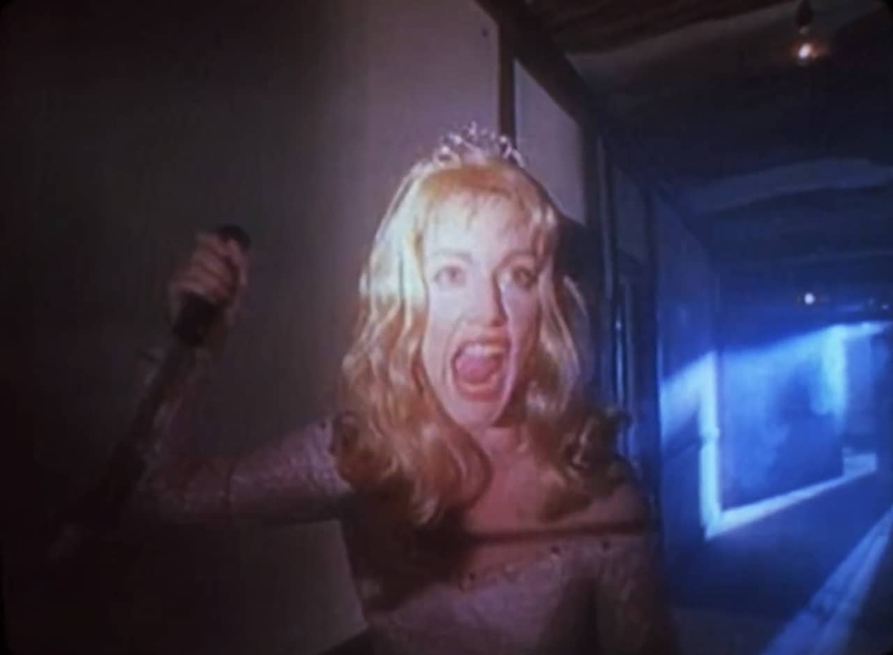 in a dark hallway, a blonde woman in a gown and tiara screams and wields a knife