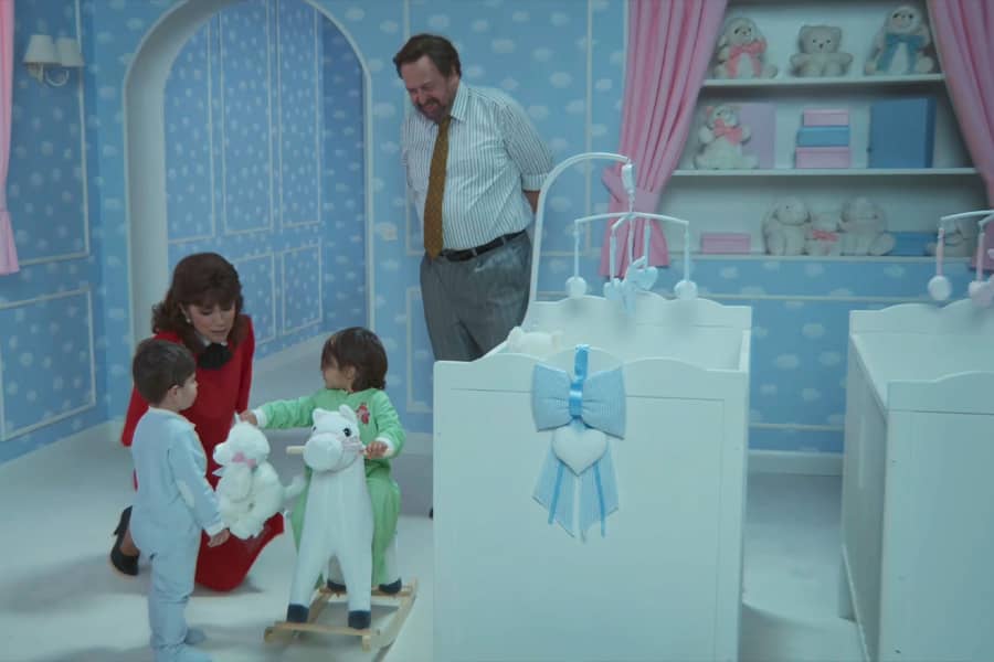 in a nursery, Pas kneels to speak with her baby twins; Minos stands nearby