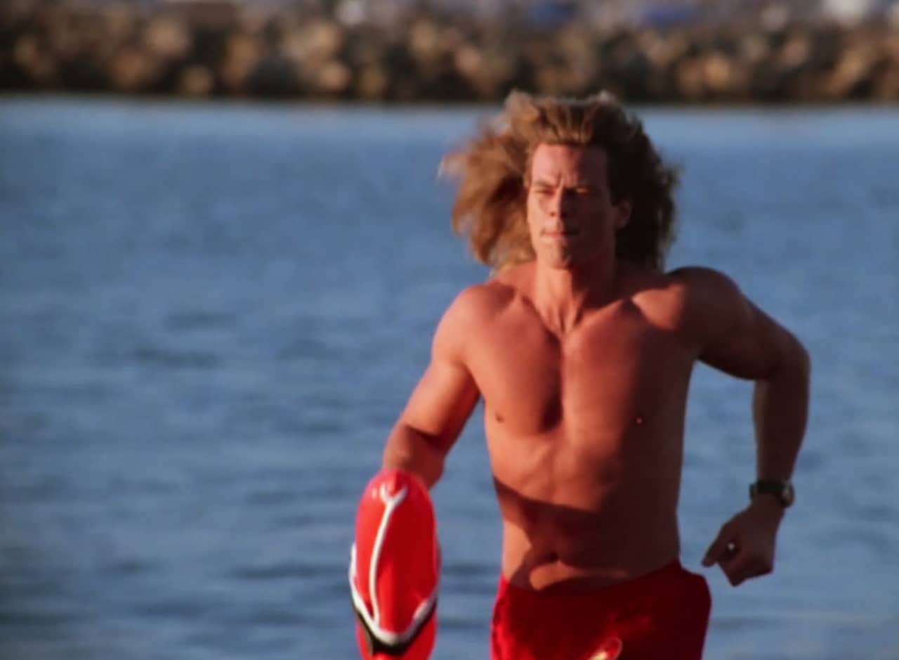 a hunky male lifeguard runs along the beach