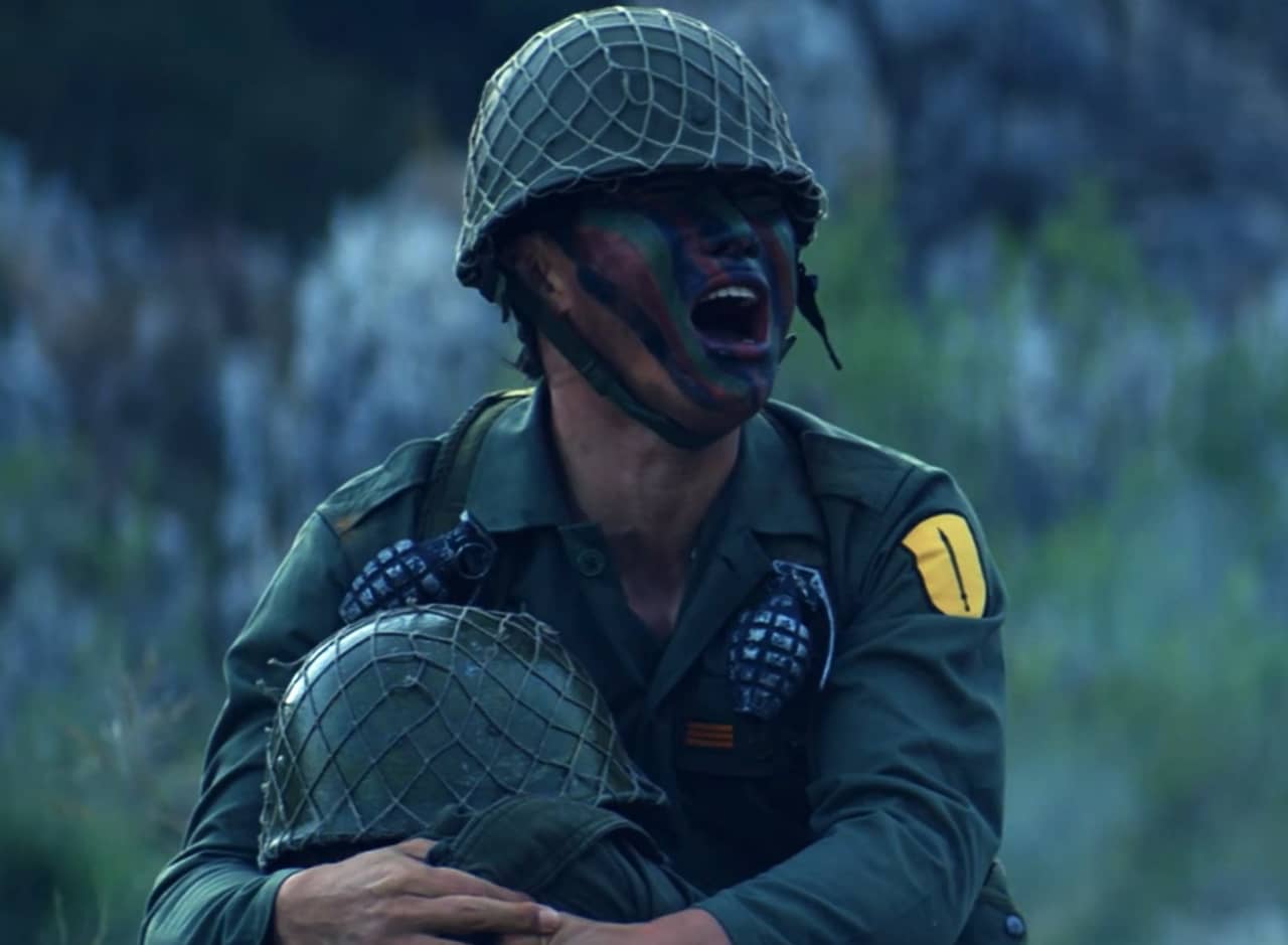 a man in military gear and war paint cries out as he holds a fallen soldier
