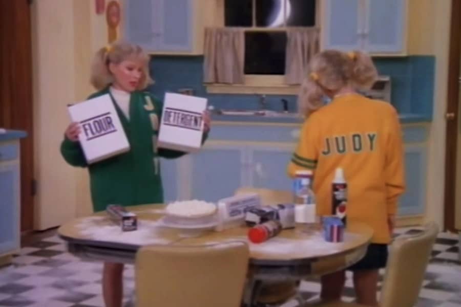 Jody and Judy baking in the kitchen, Judy is facing away from the camera, and Jody is holding two similar boxes: one is flour and the other is detergent