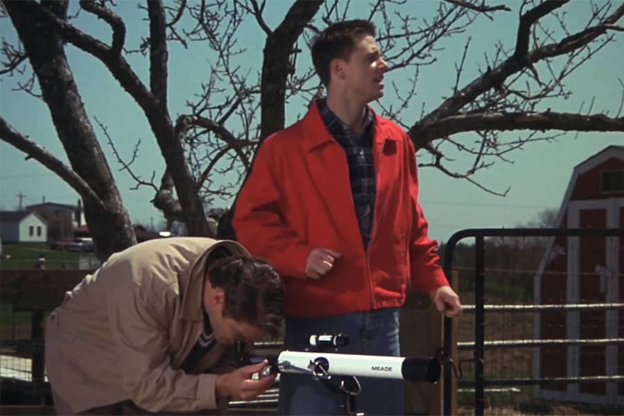 two teens waiting, one is looking through a telescope