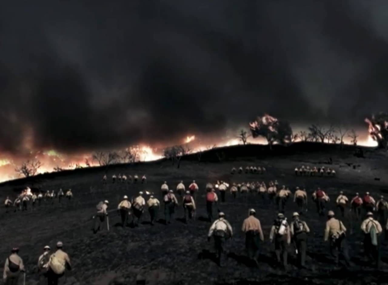 lines of firemen walk up a hill toward a raging wildfire