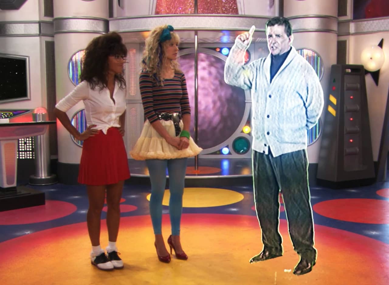 Robin Sparkles and Jessica Glitter in 90s outfits stand on their spaceship talking to a hologram Alan Thicke