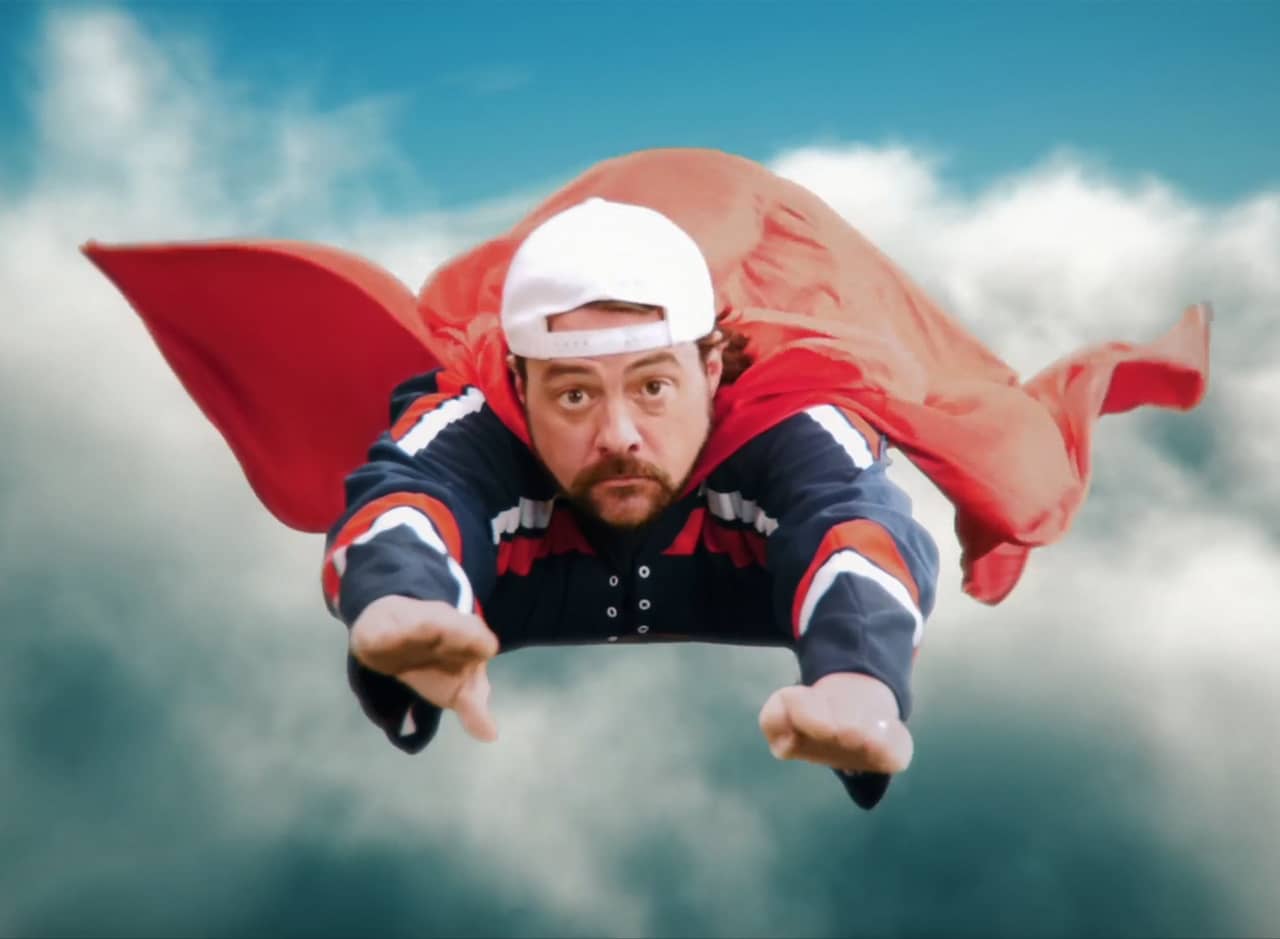 bearded man with a backwards baseball cap and a cape flies through the sky