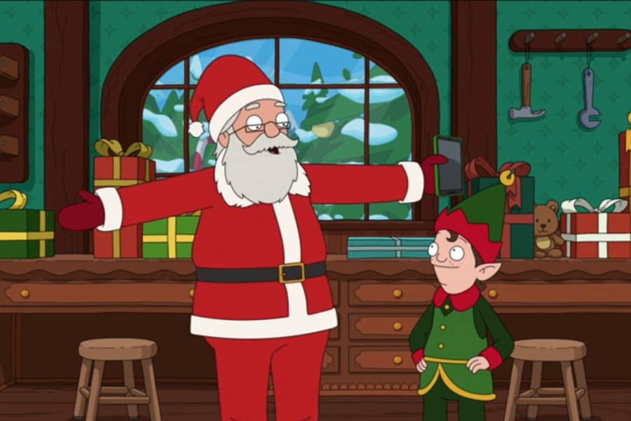 Santa holding his phone and talking with the elf in their workshop