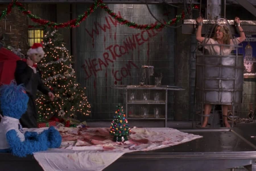 the santa baby mask man walks into a dungeon-like room with blood on the walls, a woman in a cage, and a blue muppet