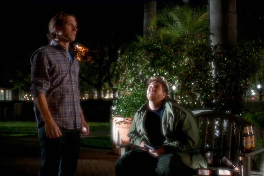 Nick stands next to a drunk man sitting on a park bench