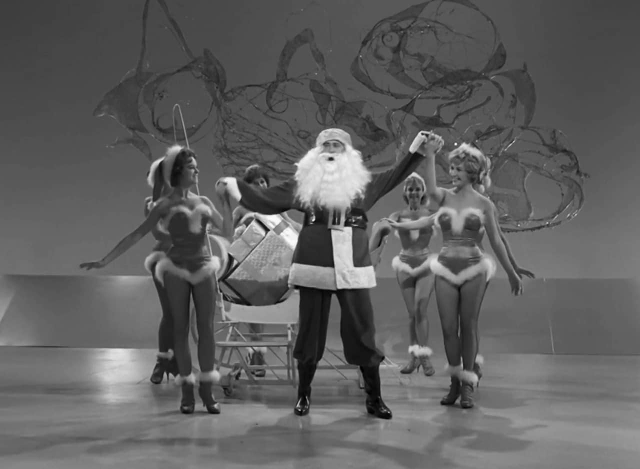 Alan Brady dressed as Santa surrounded by women in skimpy Santa outfits
