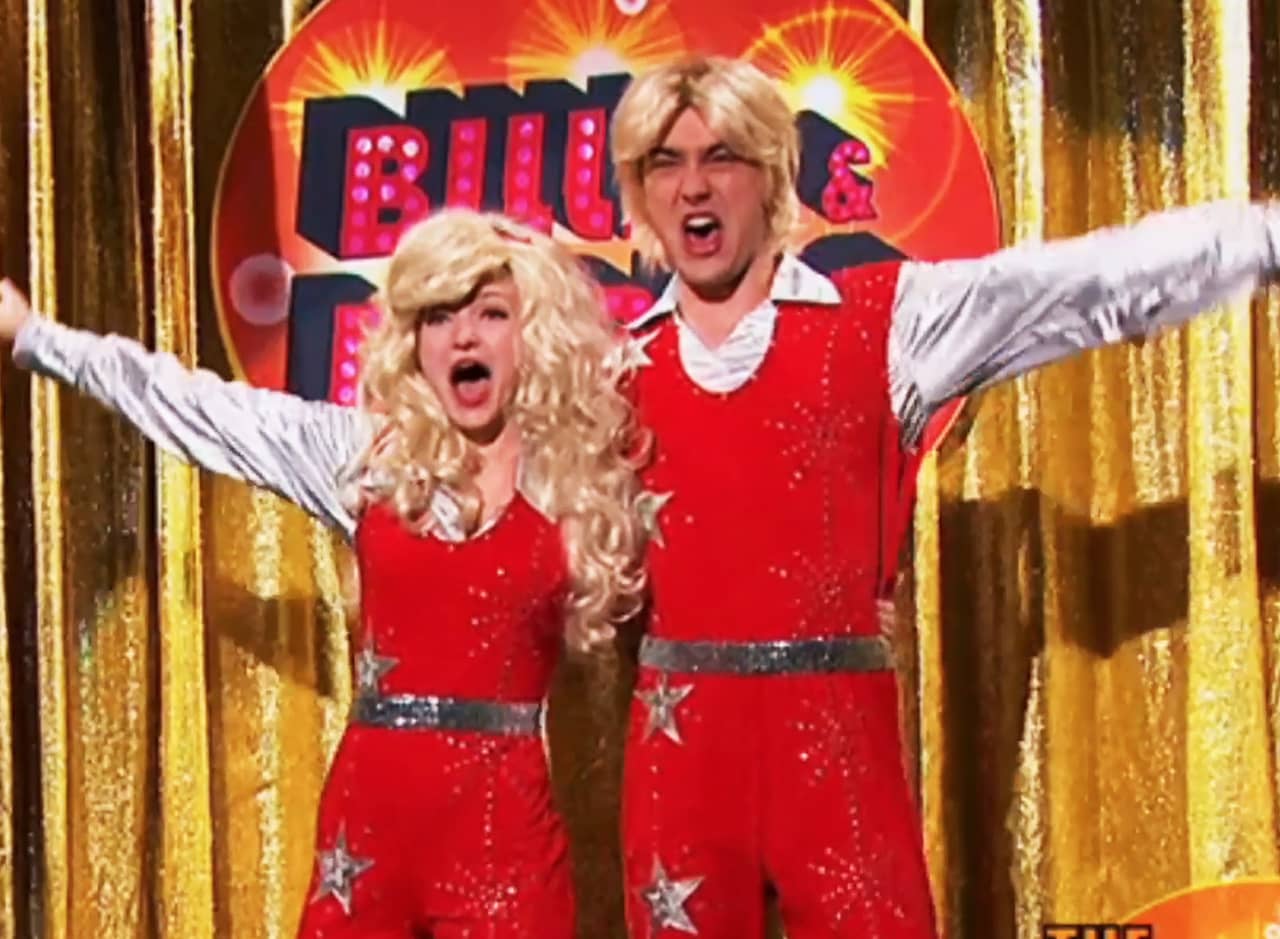 Billie, a teen boy, and Bobbie, a teen girl, both wearing blonde wigs, singing in front of a gold curtain in matching red outfits