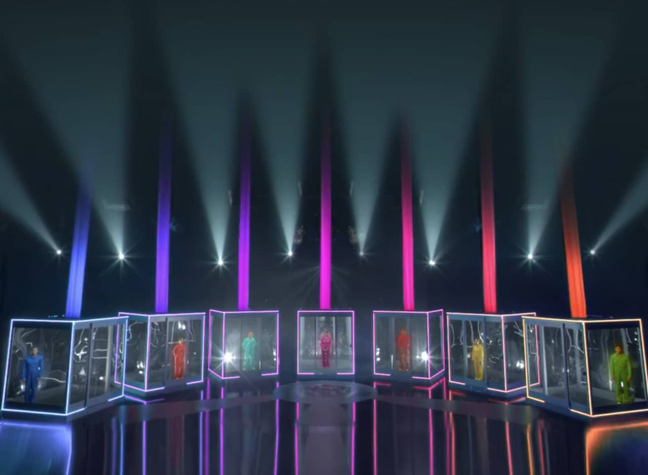 a row of large glass boxes, illuminated in different colors, each with a person inside