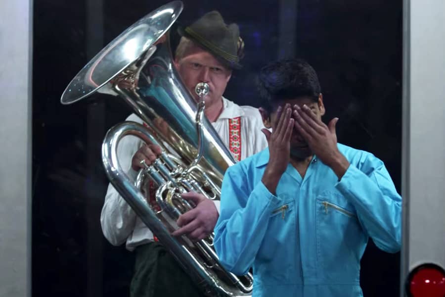 a contestant covers his face in frustration as a man in lederhosen plays a tuba behind him