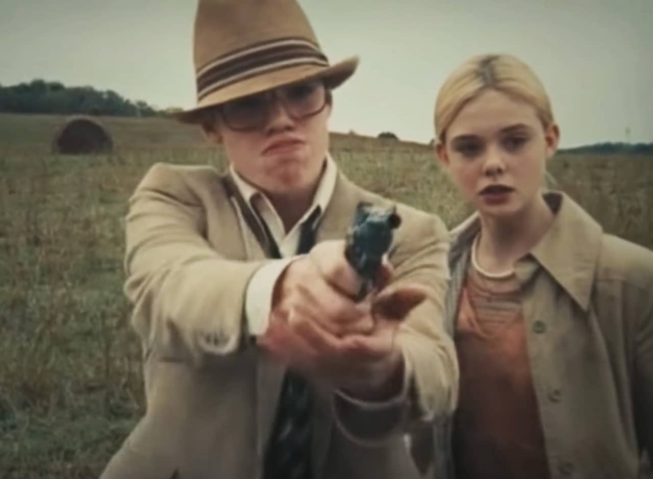 a teen boy and girl in a field, the boy raises a gun