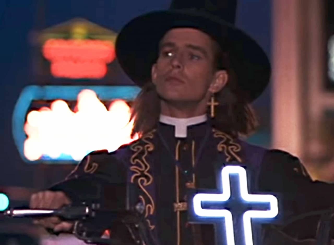 young priest with cross earring and cowboy hat, riding a motorcycle with a light-up cross on the front
