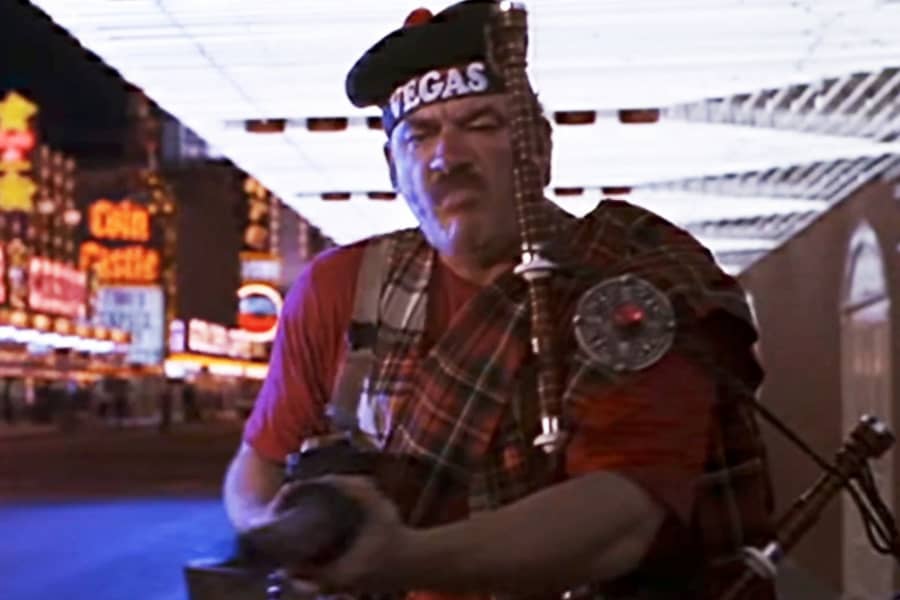 a large man in a Scottish kilt and holding bagpipes, points a gun