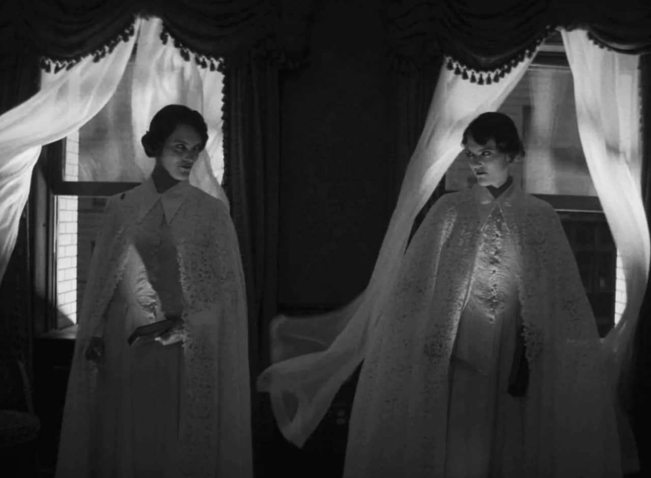 two twin women look at each other, they are wearing long lace capes and standing in front of two windows where the white curtains move in the wind