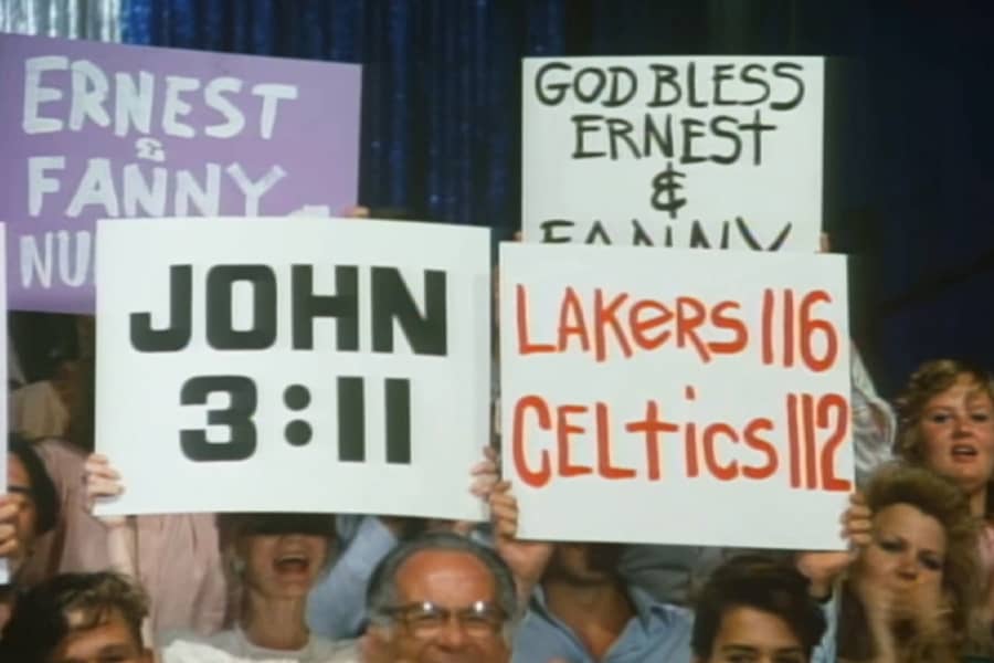 audience members holding up signs, one reads “John 3:11” and another reads “lakers 116, Celtics 112”
