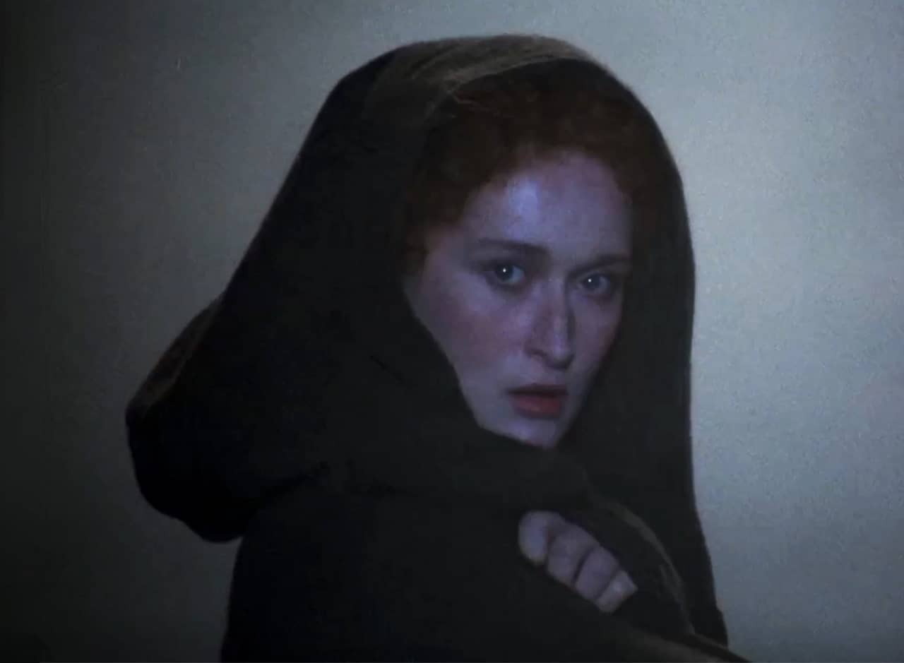 Sarah Woodruff with an intense stare from under a hooded shawl in the dark mist of the ocean