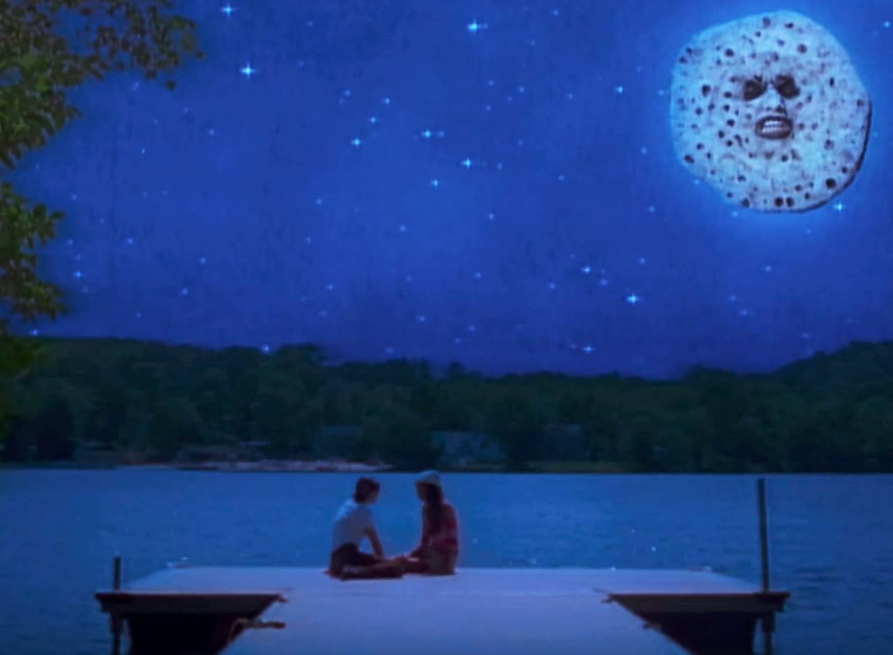 two teen girls sit on a dock by a lake in the evening; a moon with a face is in the sky