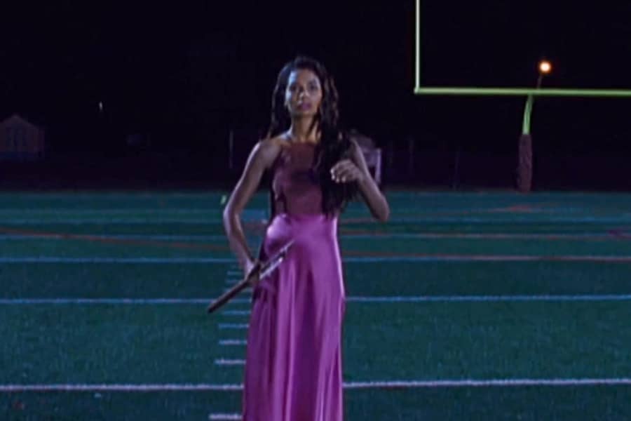 Isabel, a teen with curly hair, walks down a football field wearing a prom dress and carrying an axe