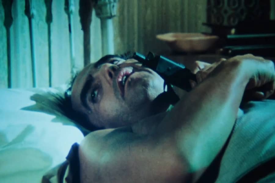 Roark lays in bed holding a gun to his face