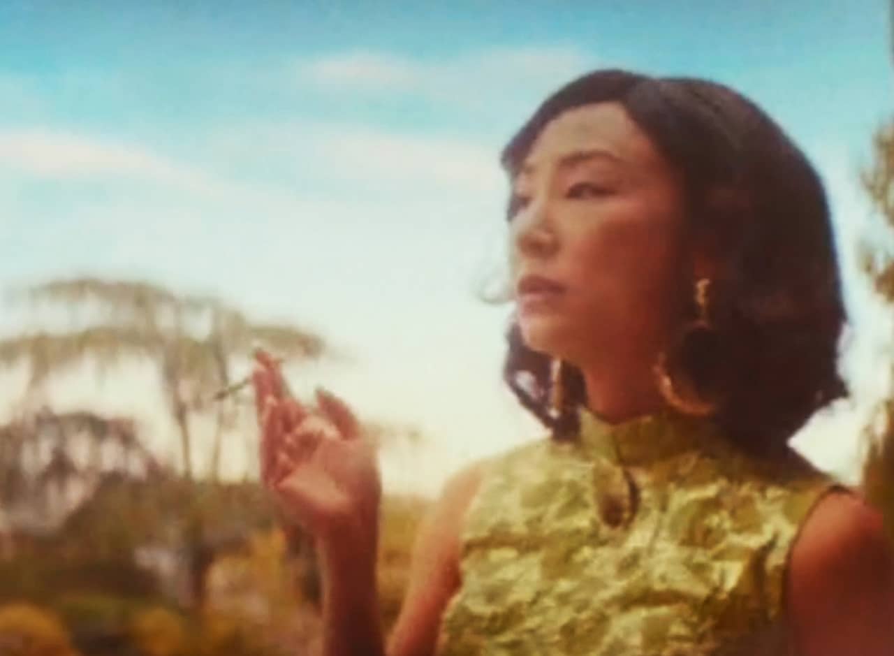 Greta Lee in a green retro strapless dress smoking a joint with blue sky and trees in the background