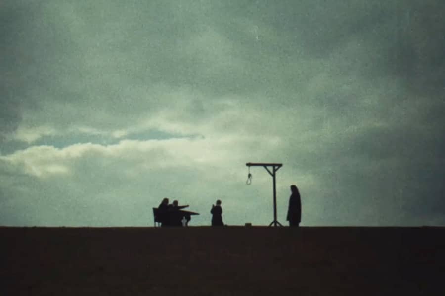 wide shot of two men at a desk, a man begging on his knees near a gallows and an executioner