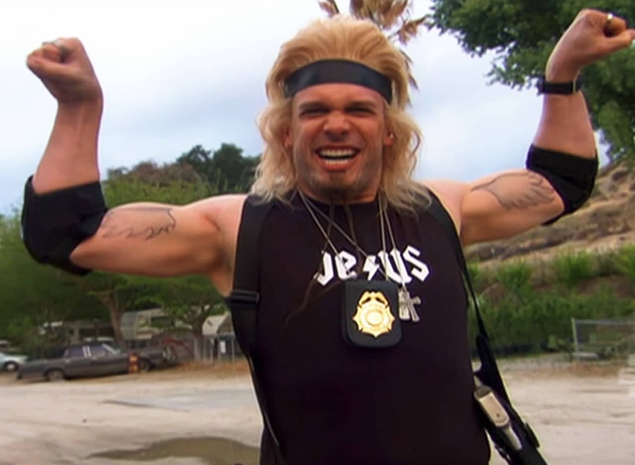 Tommy Hawk, a white man with bright blonde long hair, wearing a Jesus tank top and flexing his biceps