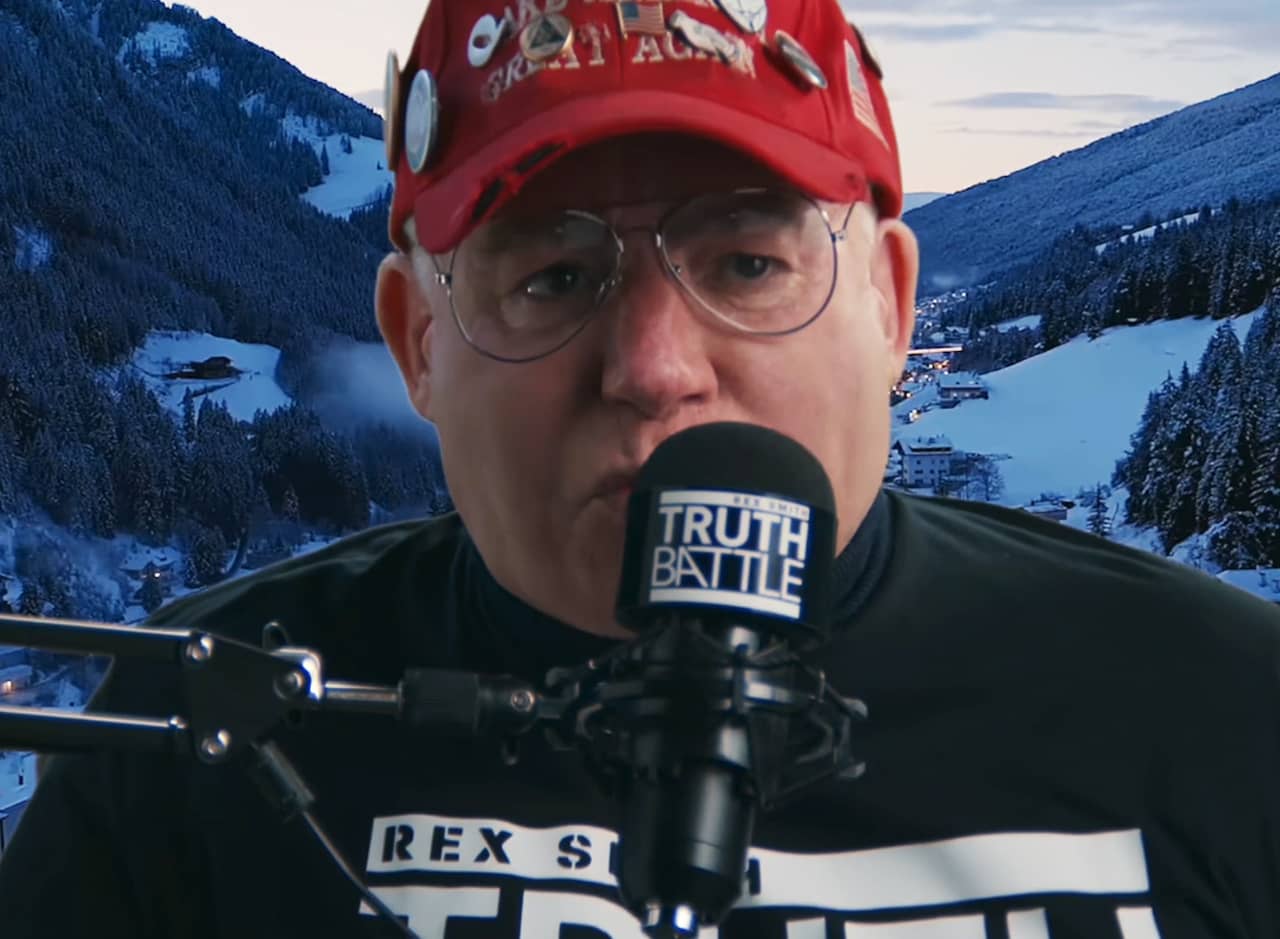 Rex Smith, an older white man with glasses and a MAGA hat, speaking into a microphone
