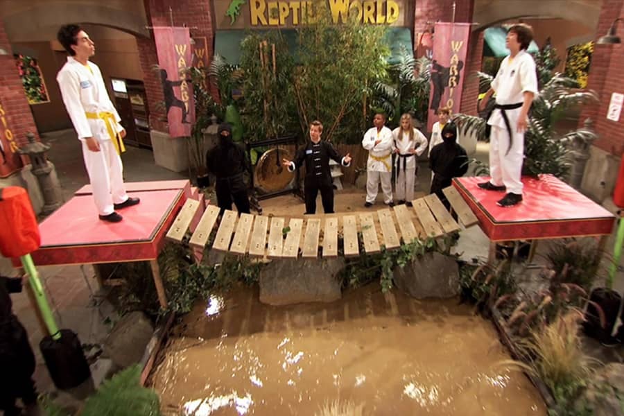two karate kids on platforms across from each other, connected by a bridge of a pool of mud