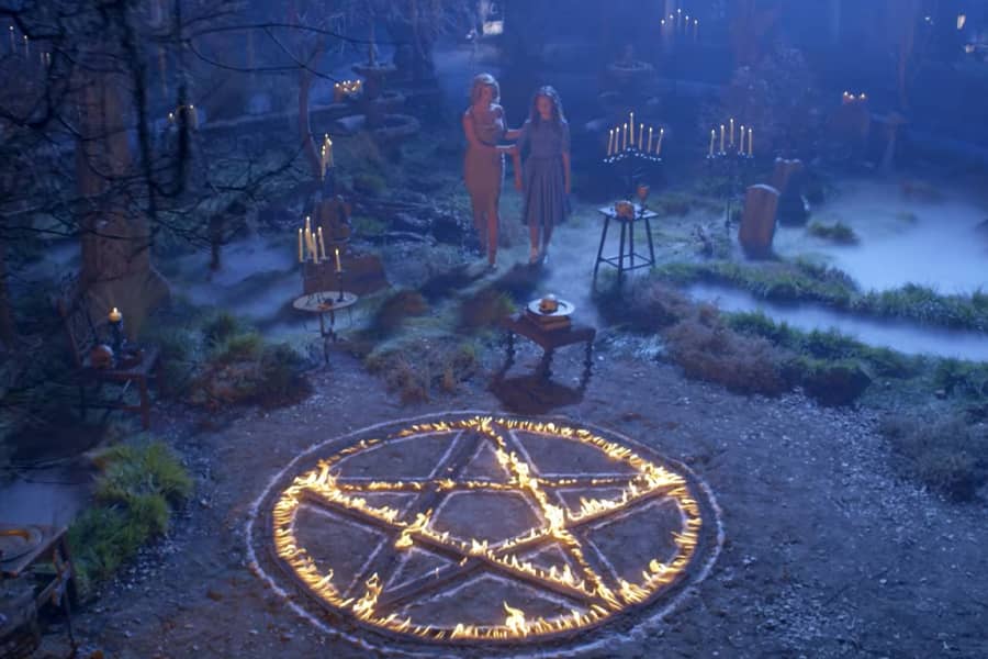 in the woods at night, two people approach a pentagram made of fire surrounded by lit candles