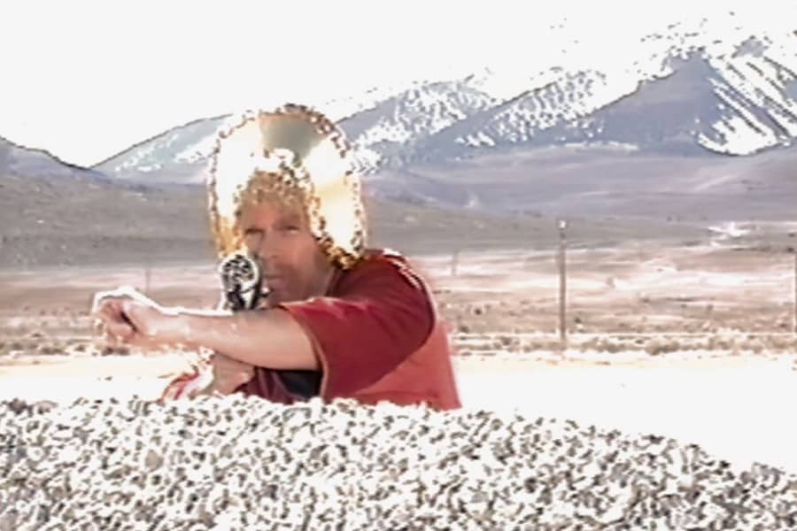 man wearing golden headdress pointing a gun from behind a rock; the shot is overexposed