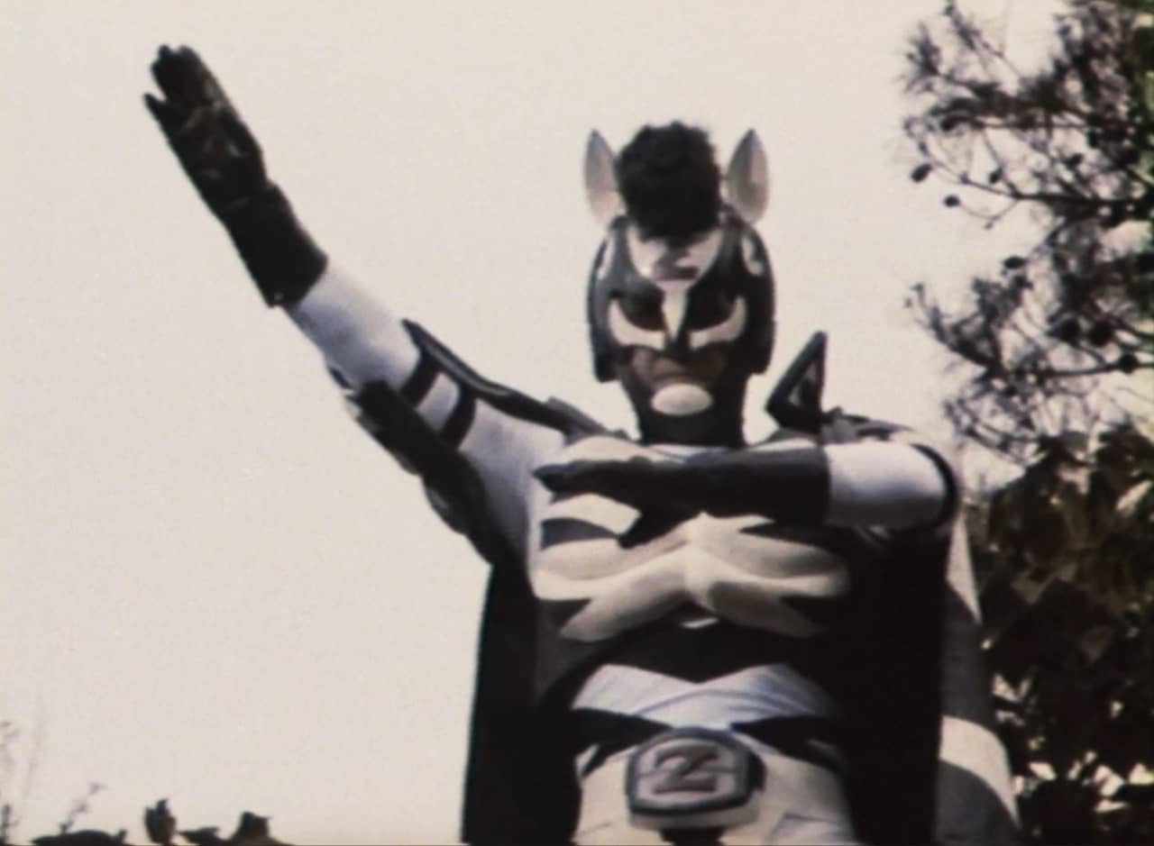 Zebraman, a superhero in a white and black striped costume, holding his gloved hand up in the air